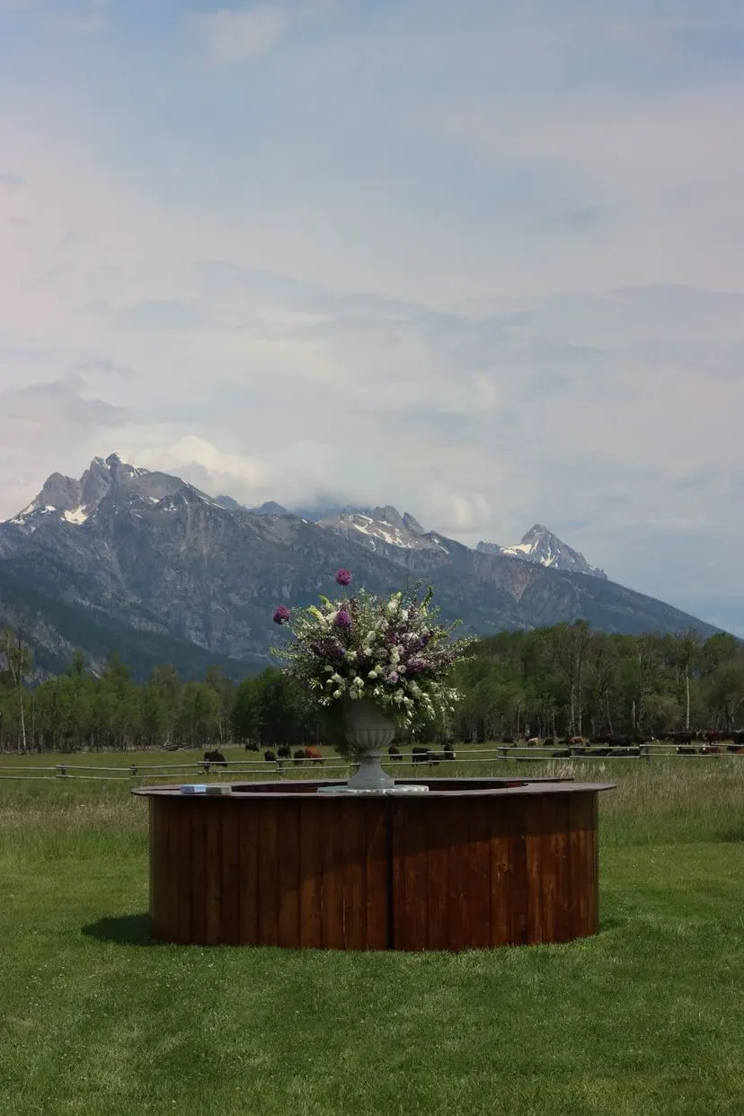Tall urn of purple and white florals on a round wood bar, with cattle and the Tetons behind it