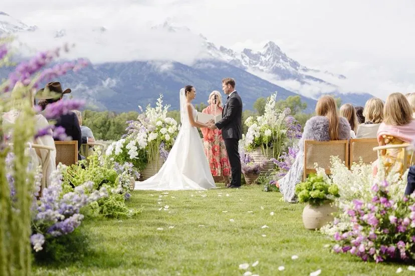 A mountain wedding ceremony with lavender ground florals and the Tetons behind the couple